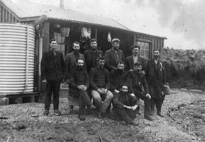 Twelve whaling men gathered outside a hut with a water tank